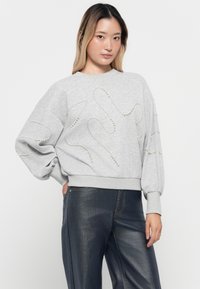 Ung kvinne iført lys grå sweatshirt med perlebeaded squiggle-mønstre og mørke vidde bukser, stående mot hvit bakgrunn.