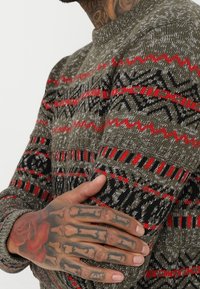 Strickpullover in olivgrün mit roten und schwarzen geometrischen Mustern. Verfügt über einen Rundhalsausschnitt und ein strukturiertes Gewebe.