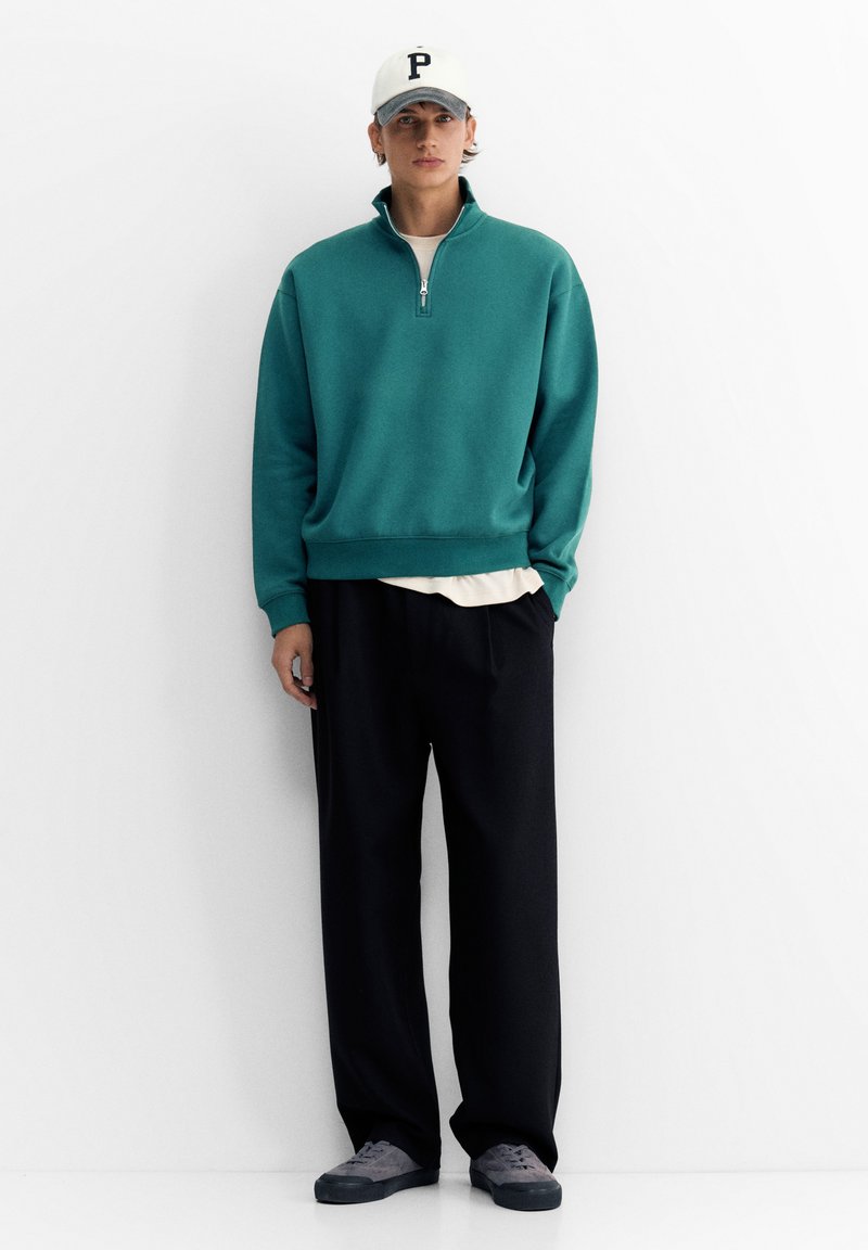 Jeune homme portant un sweat-shirt à fermeture éclair quart de couleur sarcelle, pantalon noir, baskets grises, t-shirt blanc et casquette de baseball blanche avec la lettre P, debout contre un mur blanc.