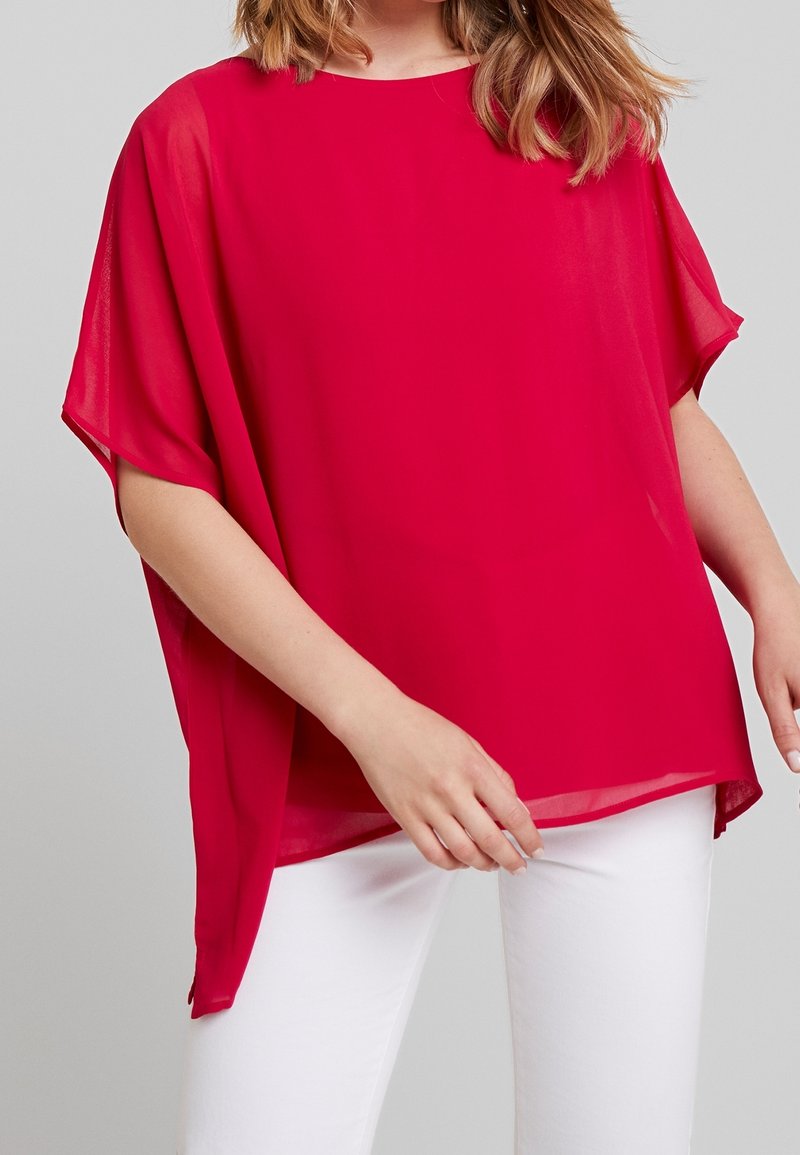 Femme portant un chemisier rouge ample à manches courtes avec un col rond et un pantalon blanc sur un fond gris uni.