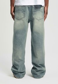 Uomo che indossa jeans azzurri larghi e sbiaditi con tasche posteriori, in piedi con le mani rilassate ai lati contro uno sfondo chiaro e semplice.