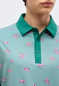 Πράσινο πουκάμισο με γιακά και μικρό καρό μοτίβο, διακοσμημένο με σκορπισμένα ροζ λουλουδένια σχέδια, κουμπωμένο στο λαιμό, που φοριέται από ένα άτομο.