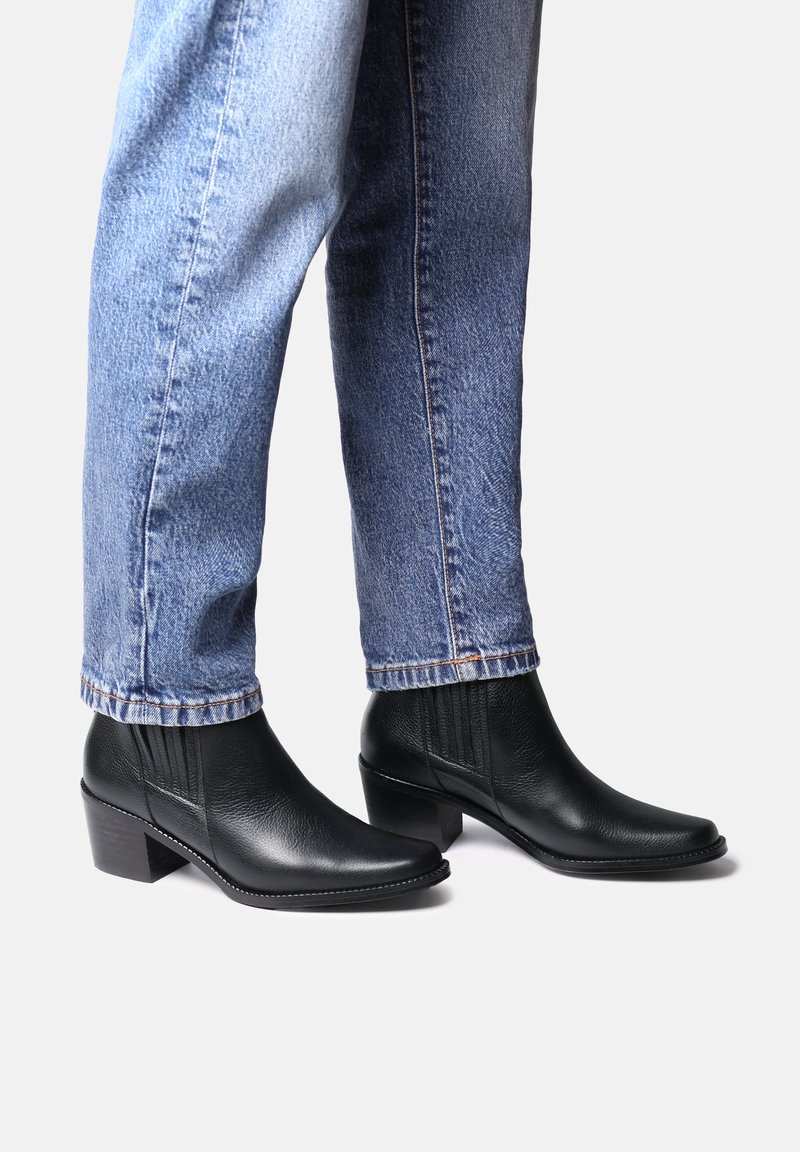 Persona che indossa stivaletti neri in pelle con tacco a blocco e jeans denim blu su uno sfondo bianco uniforme.