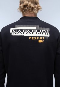 Sudadera negra con grandes gráficos en blanco y naranja con el texto "NAPAPIJRI" y "EXP.87" en la parte posterior, con un tejido suave y texturizado.