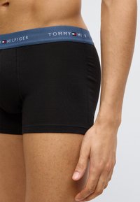 Μαύρα βαμβακερά μπαντόκ με μπλε ελαστική μέση με το λογότυπο "TOMMY HILFIGER" σε λευκό, εμφανιζόμενα από το πλάι με το χέρι τοποθετημένο στον μηρό.