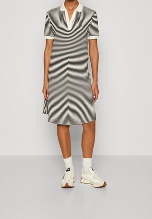 Personne portant une robe polo à rayures horizontales noires et blanches avec un col blanc, assortie à des chaussettes blanches et des baskets beige.