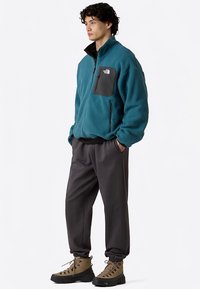 Fleece kvart-zip jakke i teal med en sort brystlomme, kombineret med grå tilspidsede bukser og brune vandrestøvler.