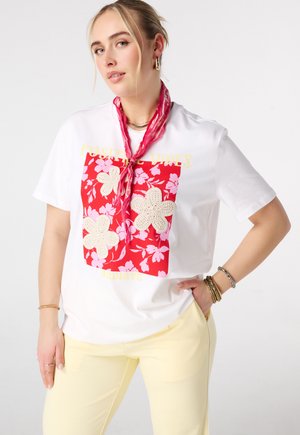 Vrouw draagt een wit t-shirt met een rood bloemmotief en gehaakte bloemenprint, gele broek, rood nekdoekje en meerdere armbanden om haar pols.