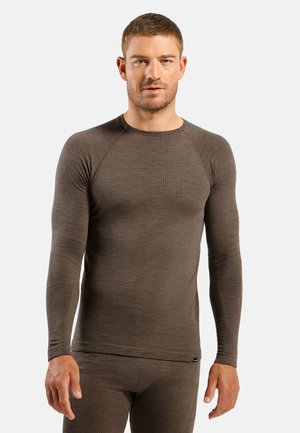 Man wearing fitted long-sleeve brown thermal top and matching pants standing against a plain light background.