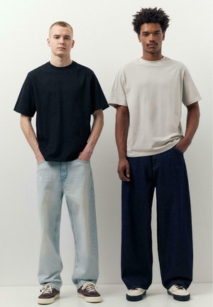 Doi bărbați stau unul lângă altul purtând tricouri simple supradimensionate și blugi largi de denim pe un fundal neutru, cu mâinile în buzunare.