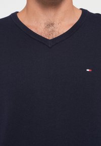 Tamno plavi pleteni pulover s V-izrezom, s malim izvezenim logotipom u crvenoj, bijeloj i plavoj boji na prsima. Mekana tekstura.