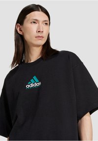 Črna bombažna oversized majica s kratkimi rokavi, ki ima teal in belo Adidasovo logo ter napis, s krožnim izrezom.