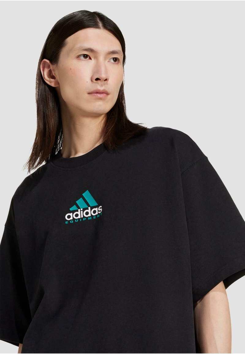 Sort bomulds oversized t-shirt med et teal og hvidt Adidas-logo og tekst, med korte ærmer og rund hals.