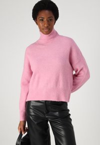 Rosa Rollkragenpullover aus weichem, flauschigem Material. Hat ein verkürztes Design und einen gerippten Saum. Getragen mit schwarzen Lederhosen.