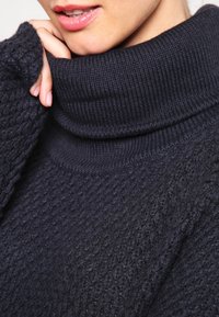 Pull à col roulé bleu marine foncé avec un motif de tricot texturé. Il dispose d'un col haut à côtes et de manches à coupe ample. Tissu doux visible.
