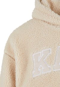 Veste beige duveteuse à capuche avec de grandes lettres blanches en contour sur la poitrine, confectionnée en matière polaire douce et texturée.
