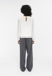 Blouse blanche avec un détail dos en trou de serrure, manches longues et coupe ample, associée à un pantalon large gris en tissu texturé.
