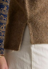 Pruun villane sviiter ribatud ääre ja külgslitsidega, mille külge on sobitatud sinine mustriline pika varrukaga särgiga, millel on ratsaspordi motiivid. Näha on valged püksid.