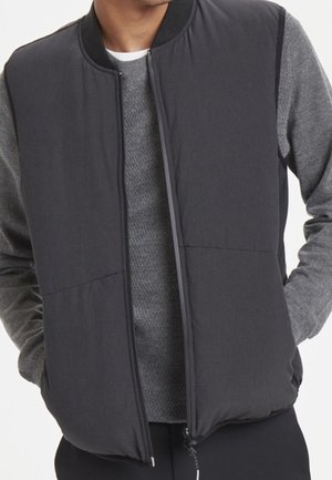 Mężczyzna w ciemnoszarej pikowanej kamizelce bez rękawów na jasnoszarym swetrze z długim rękawem, z rękami w kieszeniach kamizelki, stoi na białym tle.