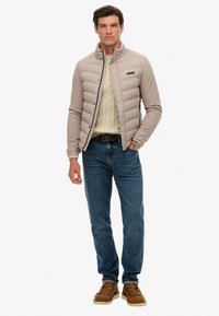 Veste matelassée beige avec un col haut, associée à un pull crème en tricot torsadé, un jean bleu et des bottes marron à lacets. Montre classique au poignet.