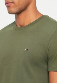 Tricou din bumbac de culoare verde măslin, cu guler rotund, mâneci scurte și un mic logo brodat cu accente roșii, albe și albastre pe piept.