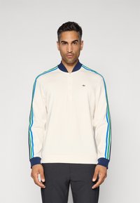 Mann som har på seg en hvit langermet genser med halv glidelås og blå, grønn og marineblå striper på ermene, kombinert med svarte bukser.