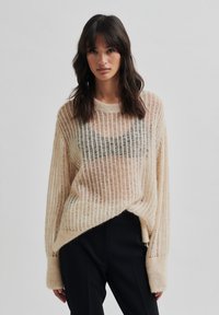 Pull en tricot beige clair avec des côtes horizontales, coupe légèrement ample, manches raglan et finition texturée. Associé à un pantalon sombre.
