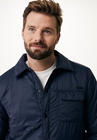 Námořnická modrá bunda na knoflíky vyrobená z lehkého materiálu. Má náprsní kapsu a hladkou texturu s jednoduchým, čistým designem.