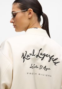 Veste couleur crème avec une texture lisse, ornée d'un texte brodé noir au dos : "Karl Lagerfeld La Côte D'Azur RIVIERA FRANÇAISE."