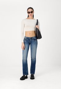 White long-sleeve crop top, blue denim jeans, and black shoes. The model holds a black shoulder bag and wears dark sunglasses.