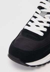 Čierne tenisky s kombináciou semišu a hladkej tkaniny. Biele šnúrky a štruktúrovaná gumová podrážka s jemným brandingom.