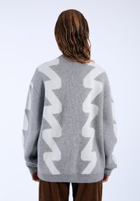 Gray knit sweater featuring a zigzag pattern in light gray, with dropped shoulders and ribbed cuffs. Soft texture and oversized fit.