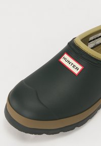 Scarpa nera in gomma slip-on con una finitura testurizzata, un interno color crema e un'etichetta rossa e bianca "HUNTER" sul lato esterno.
