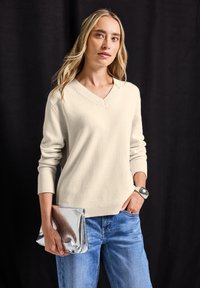 Beige v-halstrui met een gestructureerde brei, geribbelde manchetten en zoom. Gecombineerd met lichtblauwe jeans en een zilver metallic clutch.