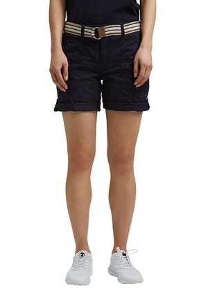 Personne portant un short noir à ourlet roulé avec des poches avant et une ceinture en tissu rayé, associée à des baskets blanches, debout.