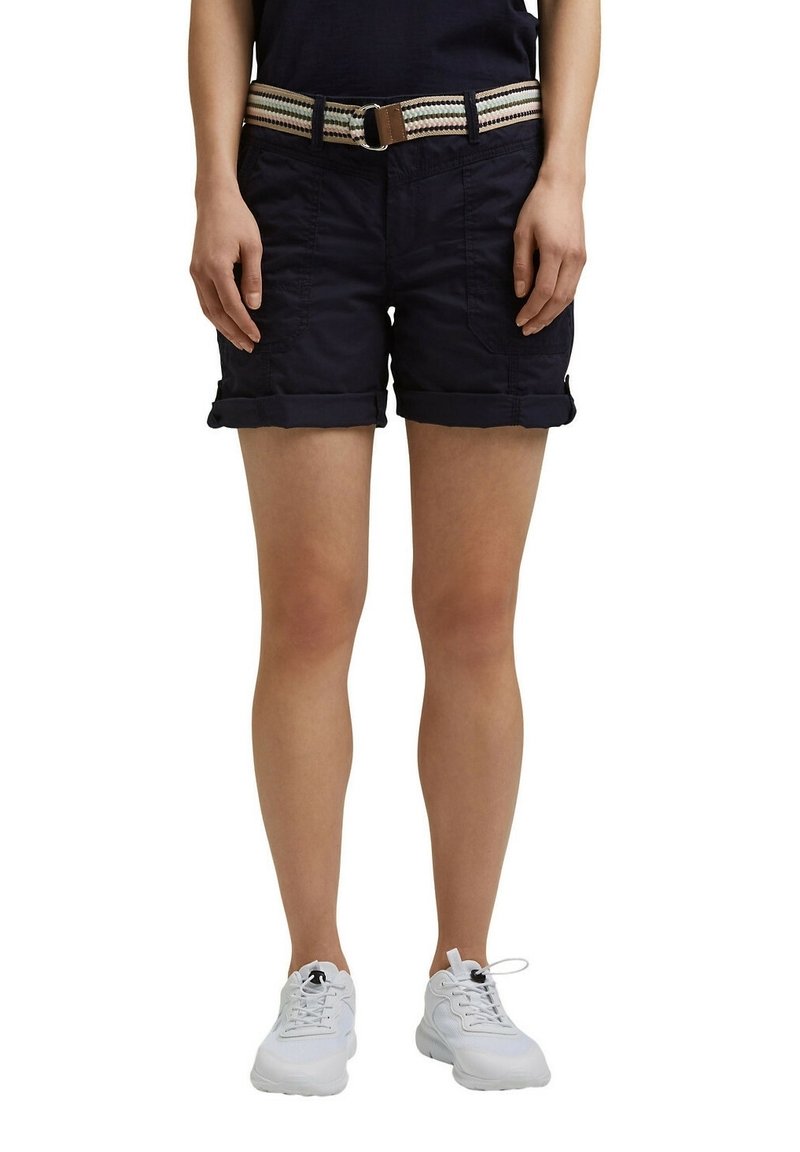Personne portant un short noir à ourlet roulé avec des poches avant et une ceinture en tissu rayé, associée à des baskets blanches, debout.