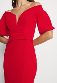 Femme portant une robe rouge décolletée, avec des manches bouffantes et un décolleté en V profond, laissant apparaître une partie de son cou et ses boucles d'oreilles.