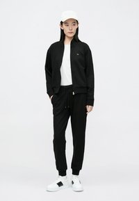 Black zip-up jacket, white t-shirt, and black joggers with drawstring. White sneakers with black accents. Model wearing a white cap.