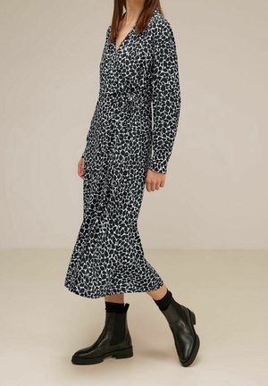 Femme portant une robe midi à manches longues, à motifs noirs et blancs avec une ceinture à la taille, associée à des bottines noires et des chaussettes noires.