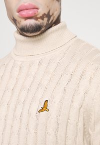 Beige gestrickter Pullover mit einem gerippten Rollkragen, der ein Zopfmuster aufweist und ein kleines gelbes Vogel-Emblem auf der Brust hat.