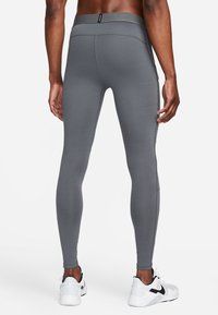 Vista trasera de una persona que lleva leggings ajustados grises y zapatillas Nike blancas sobre un fondo blanco.