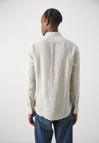 Camisa de lino beige claro con cuello, mangas largas y puños abotonados. Con dobladillo ligeramente curvado, combinada con jeans de denim azules.