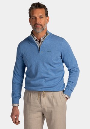 Man met kort grijs haar en baard, gekleed in een lichtblauwe trui met een kwartritssluiting over een overhemd met kraag en beige broek, staand met één hand in zijn zak.