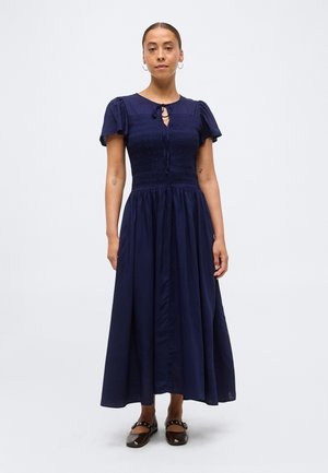 Femme debout portant une robe midi bleu marine avec des manches courtes bouffantes, un corsage froncé, un col en forme de goutte d'eau, et des chaussures Mary Jane noires.