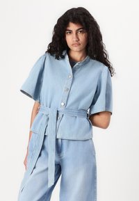 Light blue denim shirt with short puff sleeves, round collar, front buttons, and a belted waist. Soft texture and relaxed fit.