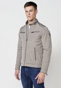 Chaqueta gris claro con cremallera y cuello alzado. Presenta dos bolsillos en el pecho con cremallera y un bolsillo lateral. Textura suave con un diseño discreto.