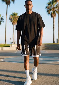 Bărbat care merge pe o teren de baschet în aer liber, purtând un tricou negru supradimensionat, pantaloni scurți negri cu dungi albe, șosete albe și adidași albi.