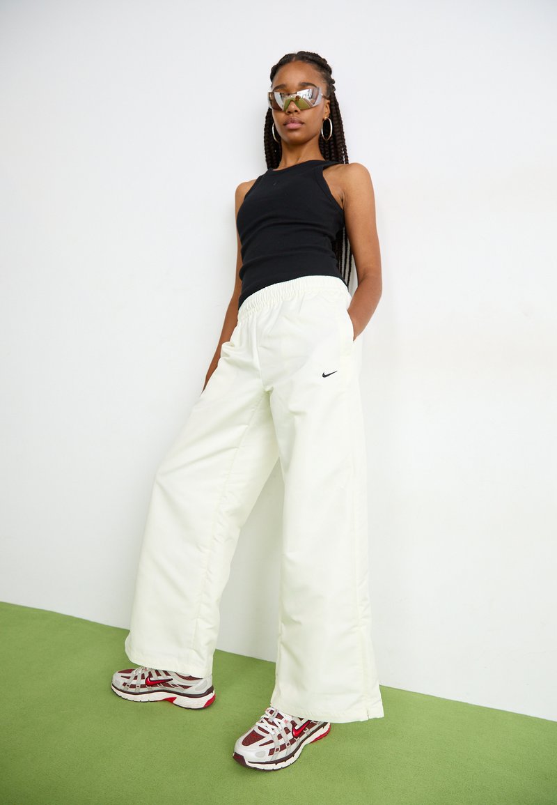 Pantalon blanc ample avec une taille élastique et un logo Nike noir. Associé à un débardeur noir et des baskets gris-rouge.
