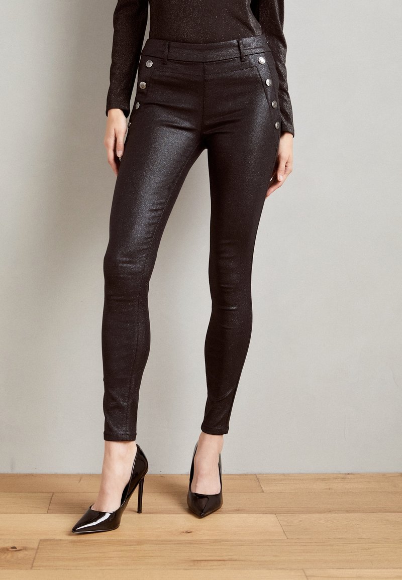 Femme portant un pantalon noir brillant et moulant avec des boutons argentés sur les côtés et des chaussures noires à talons hauts et bouts pointus, debout sur un sol en bois.