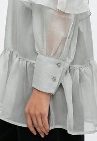 Höyhenenkevyt vaaleanharmaa pusero, jossa on röyhelöreunus ja pitkät puffihihat. Siinä on napillinen kalverin yksityiskohta ja pehmeä, laskeutuva kangaspinta.